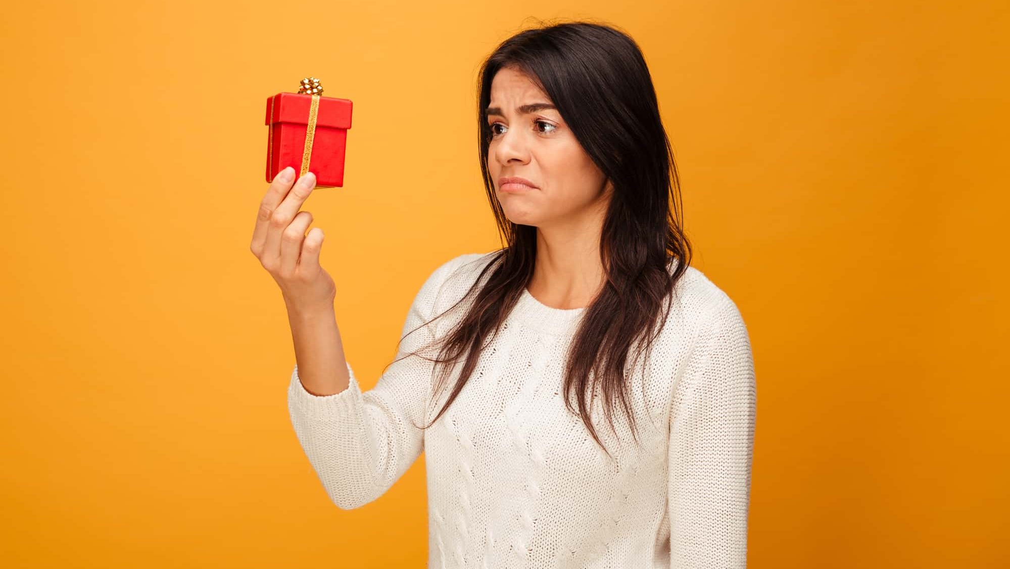Portrait of a sad young woman holding small gift box isolated over yellow background. Sad. Upset.