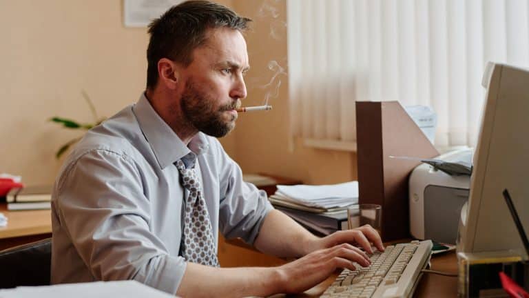 Man sitting at desk in office, smoking cigarette while typing on keyboard. Papers and office supplies scattered around giving busy work environment impression