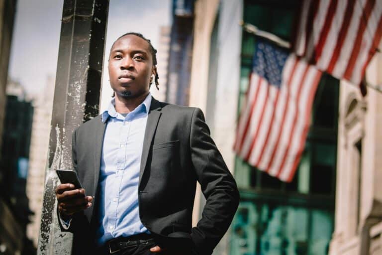 confident-black-businessman-with-smartphone-against-facade-of-building-with-us-flags