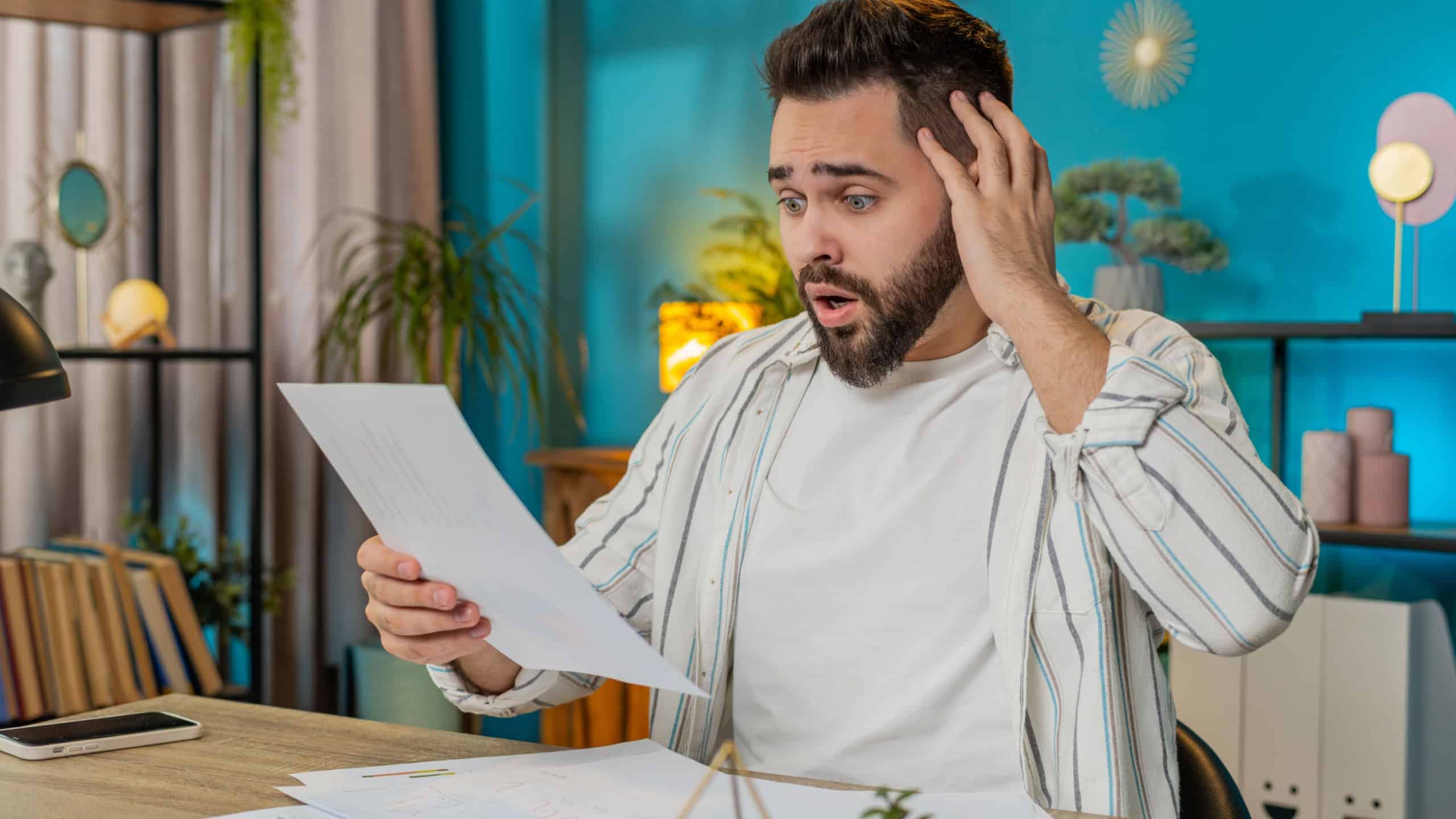 Frustrated upset shocked business man works at office desk looking at bills paperwork reports documents gets angry frustrated, stress bankruptcy financial problem. Stresses freelancer at home