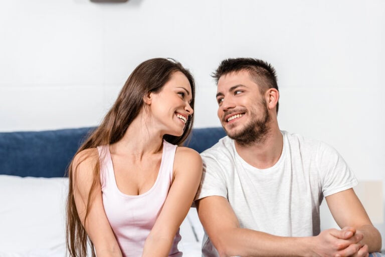 Smiling girlfriend and boyfriend in pajamas sitting on bed in bedroom and looking at each other
