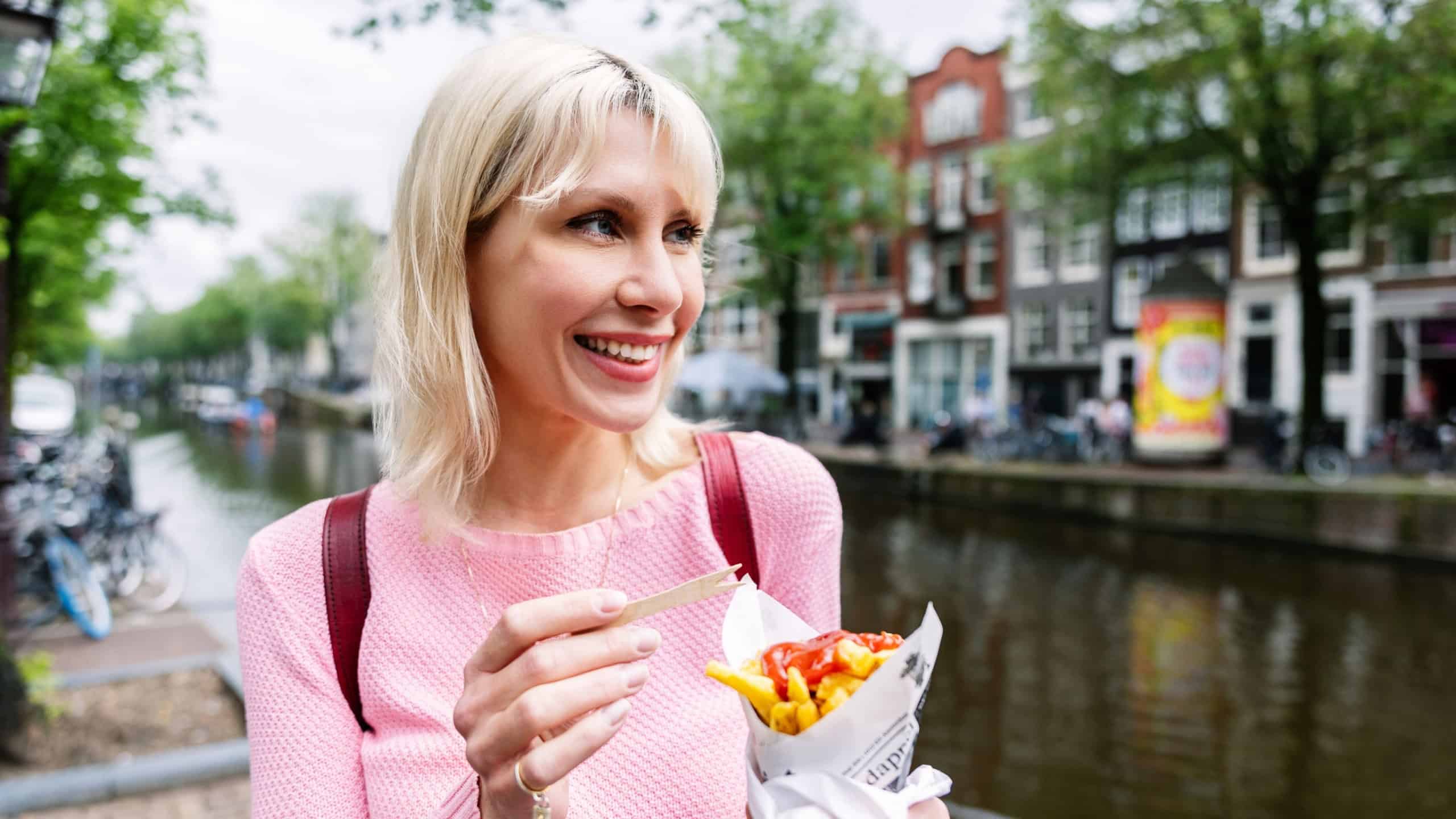 Happy young tourist woman eating fries while enjoying summer vacation in Amsterdam, Netherlands. Food and travel lifestyle concept.
