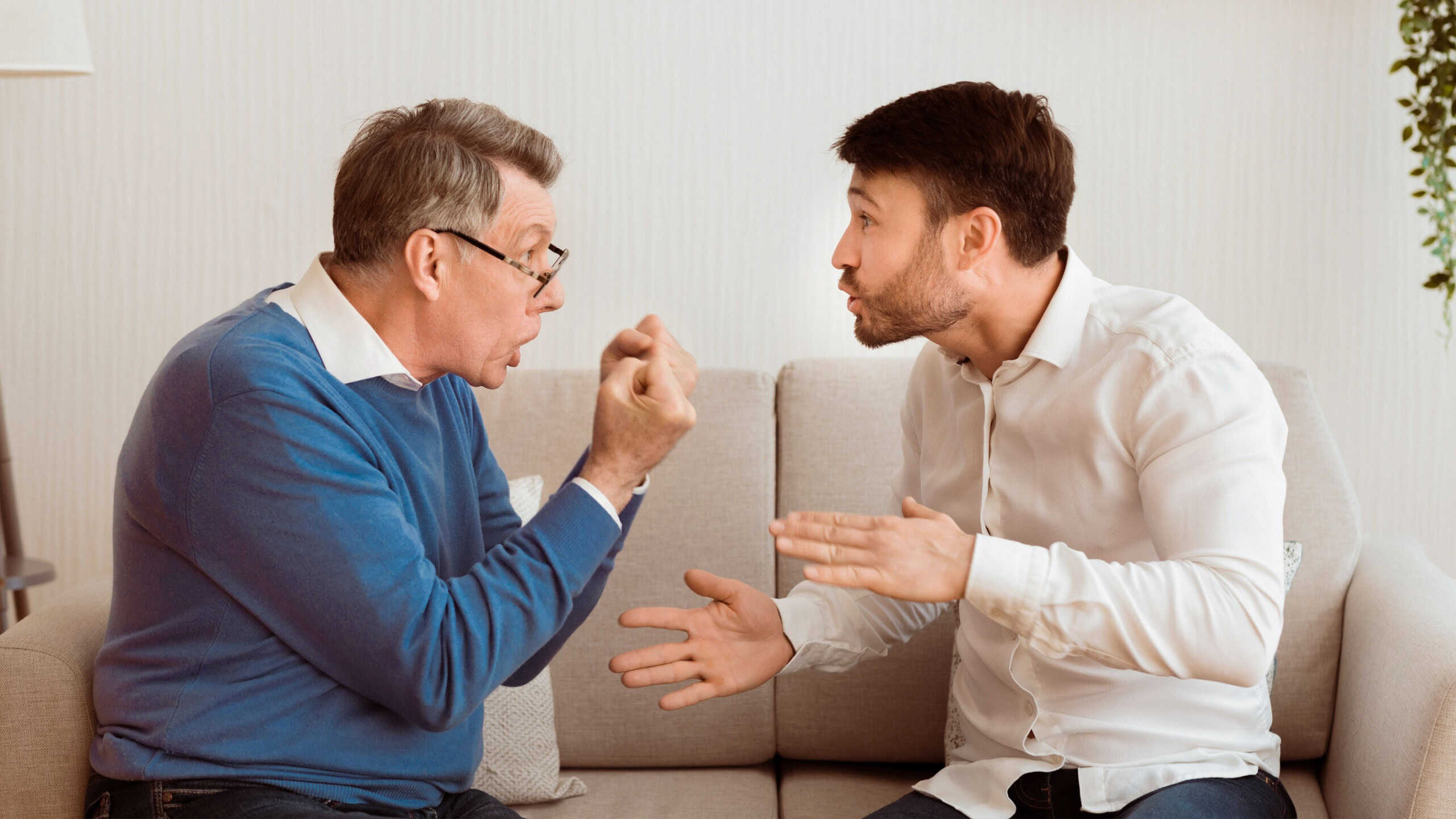 Father And Son Quarrel. Aggressive Senior Man Having Argument With Adult Son Sitting On Couch Indoor. Panorama, Selective Focus