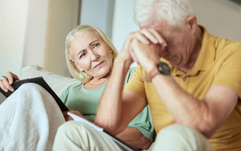 Unhappy elderly couple sitting on a sofa together and looking stressed. Senior caucasian man and woman looking worried about their future while looking at paperwork and their debt.