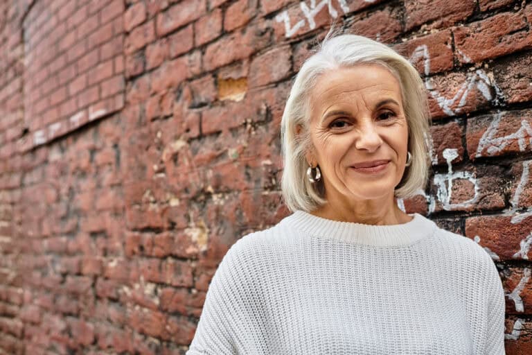 A charming older woman with silver hair leans against a brick wall