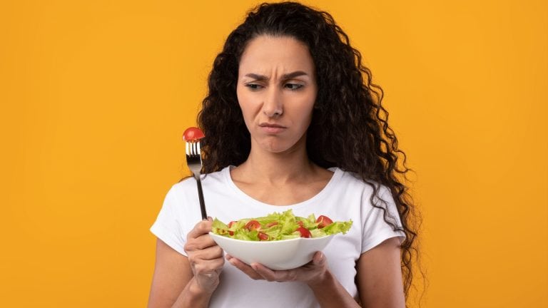 Lack Of Appetite. Portrait Of Unhappy Lady Holding Bowl Plate With Vegetable Salad And Looking At Fork Thinking About Meal Standing Isolated On Yellow Orange Studio Background. Loss Of Taste And Smell