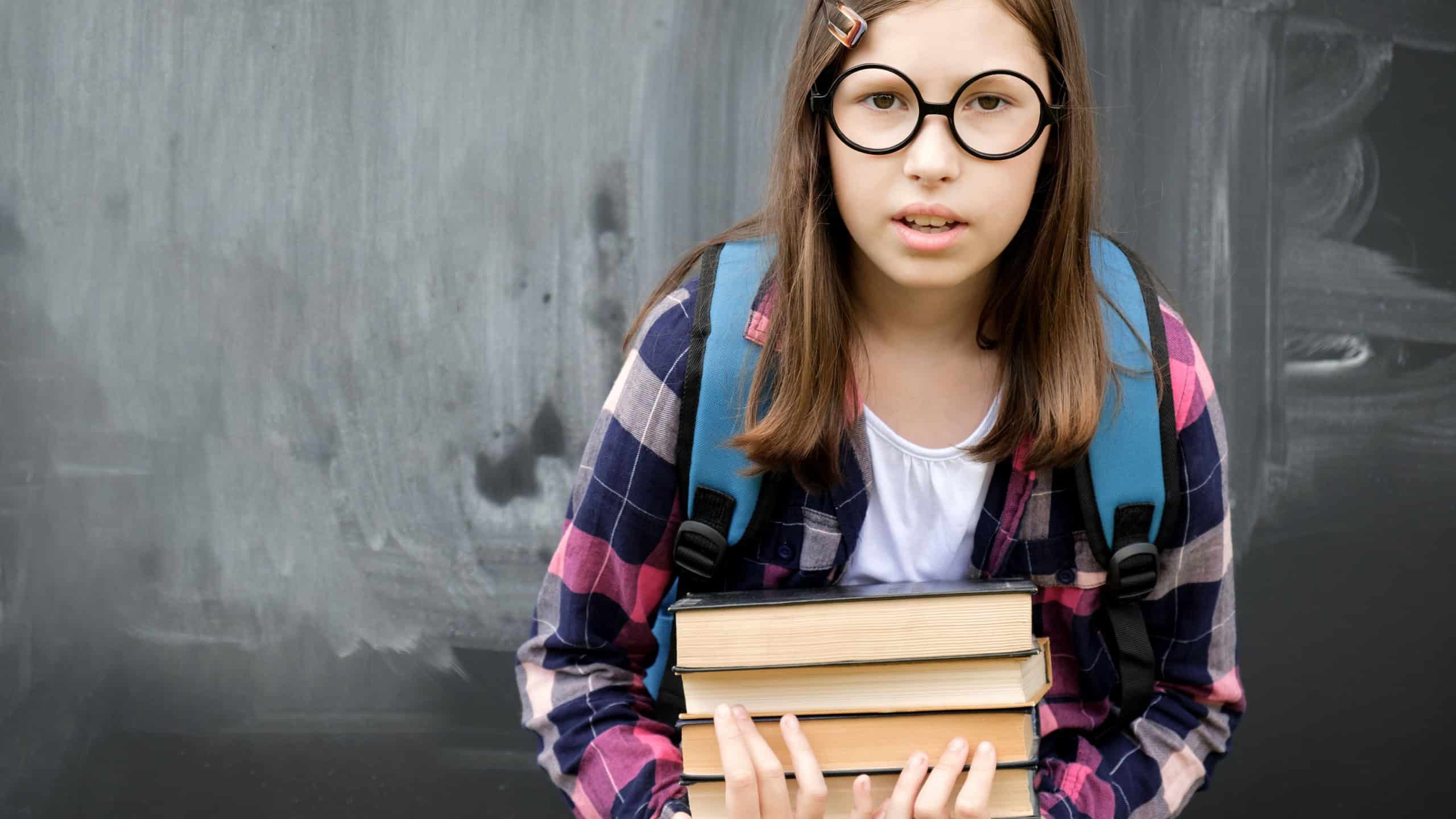 Teen girl little student holding pile of heavy books on chalkboard background. Teenage girl in shirt, glasses with blue backpack and overweight stack of textbooks. Education, school and people concept