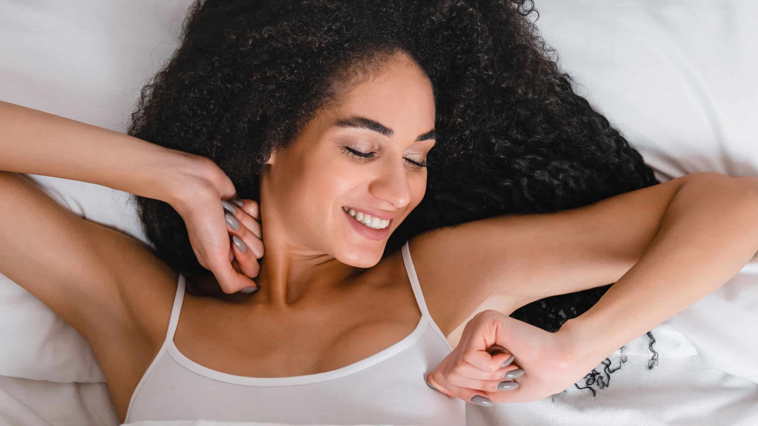 Young african happy cheerful woman in white pajamas waking up after sleeping, stretching and full of energy to start new day in the morning in the hotel room