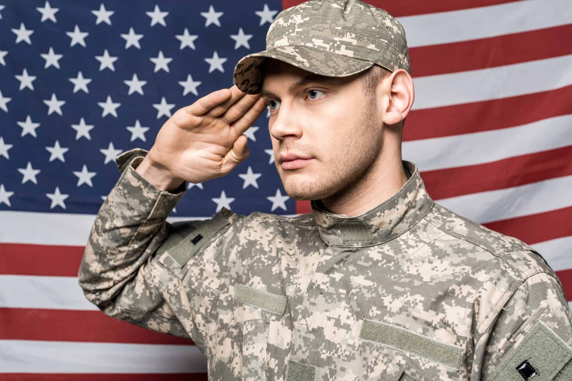 Handsome soldier in military uniform and cap giving salute near flag of america
