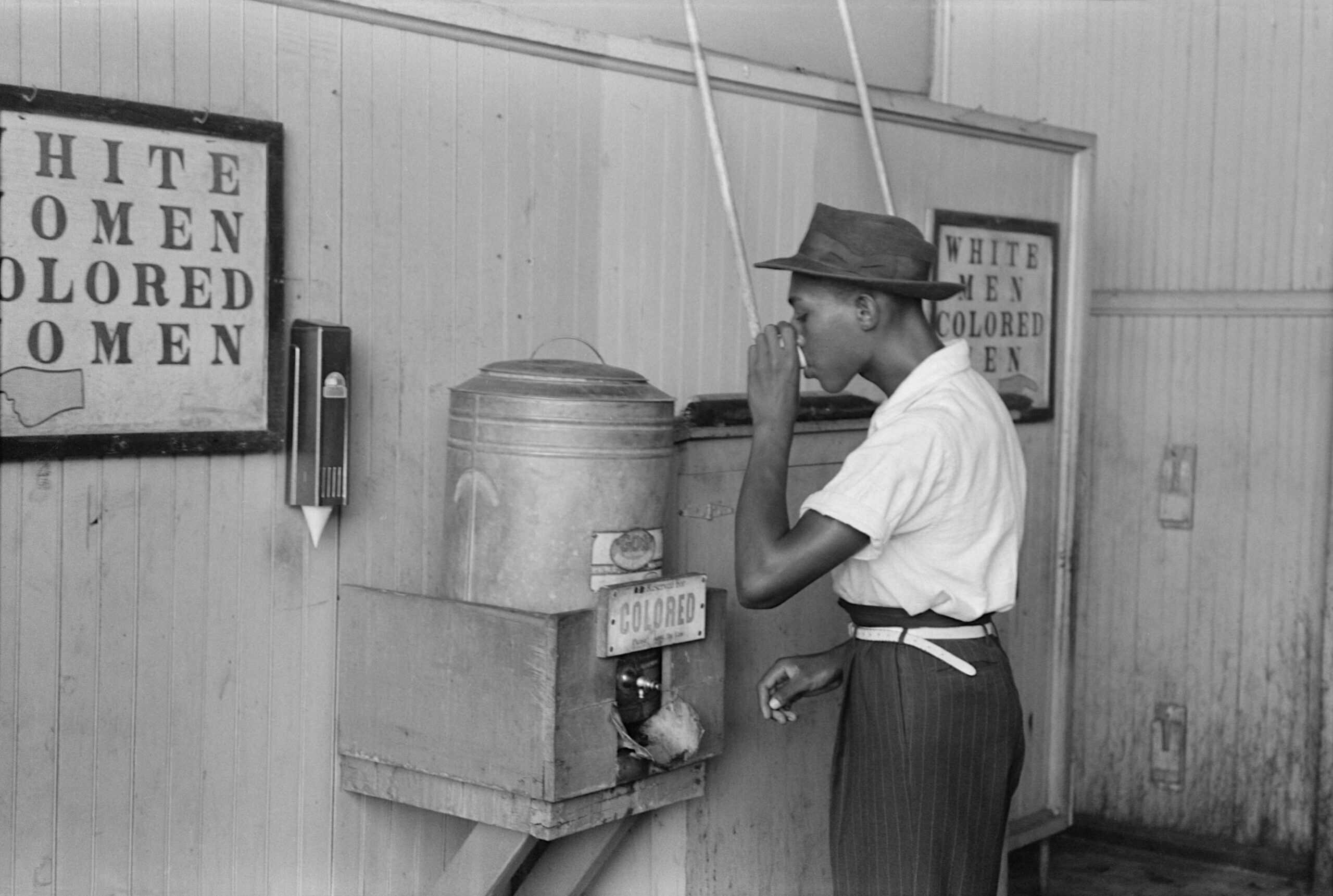 'A person of color drinking at "Colored" water cooler in streetcar terminal, racism, xenophobia