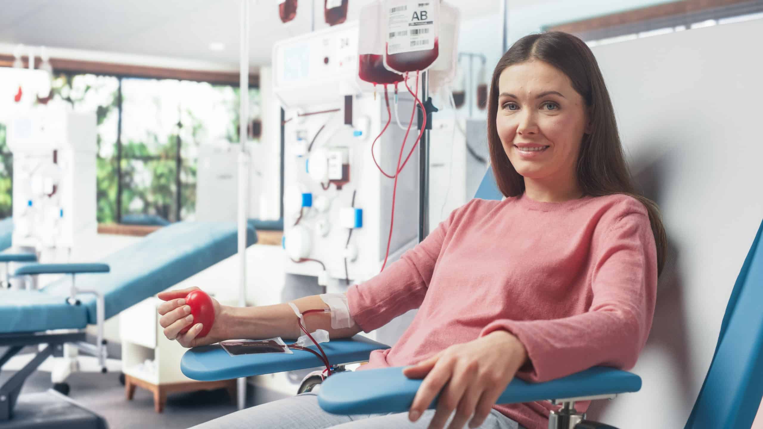 Caucasian Woman Donating Blood For People In Need In Hospital. Female Donor Squeezing Heart Shaped Ball To Pump Blood. Looking Into Camera And Smiling. Donation Center For Victims Of Accidents.