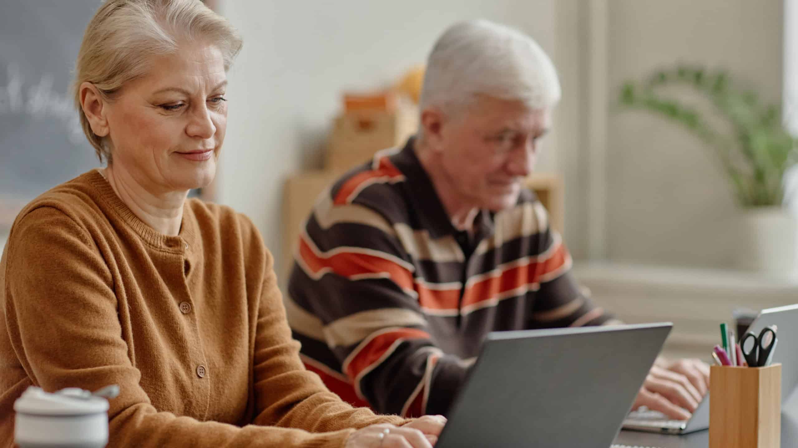Senior Caucasian woman using laptop while sitting next to senior Caucasian man working on laptop in classroom setting, both focused on learning technology skills in educational environment