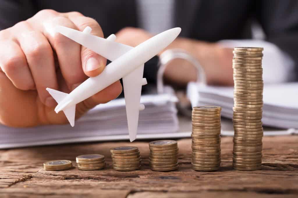 Close-up Of A Businessperson's Hand Flying Aeroplane Over Stacked Coins At Workplace