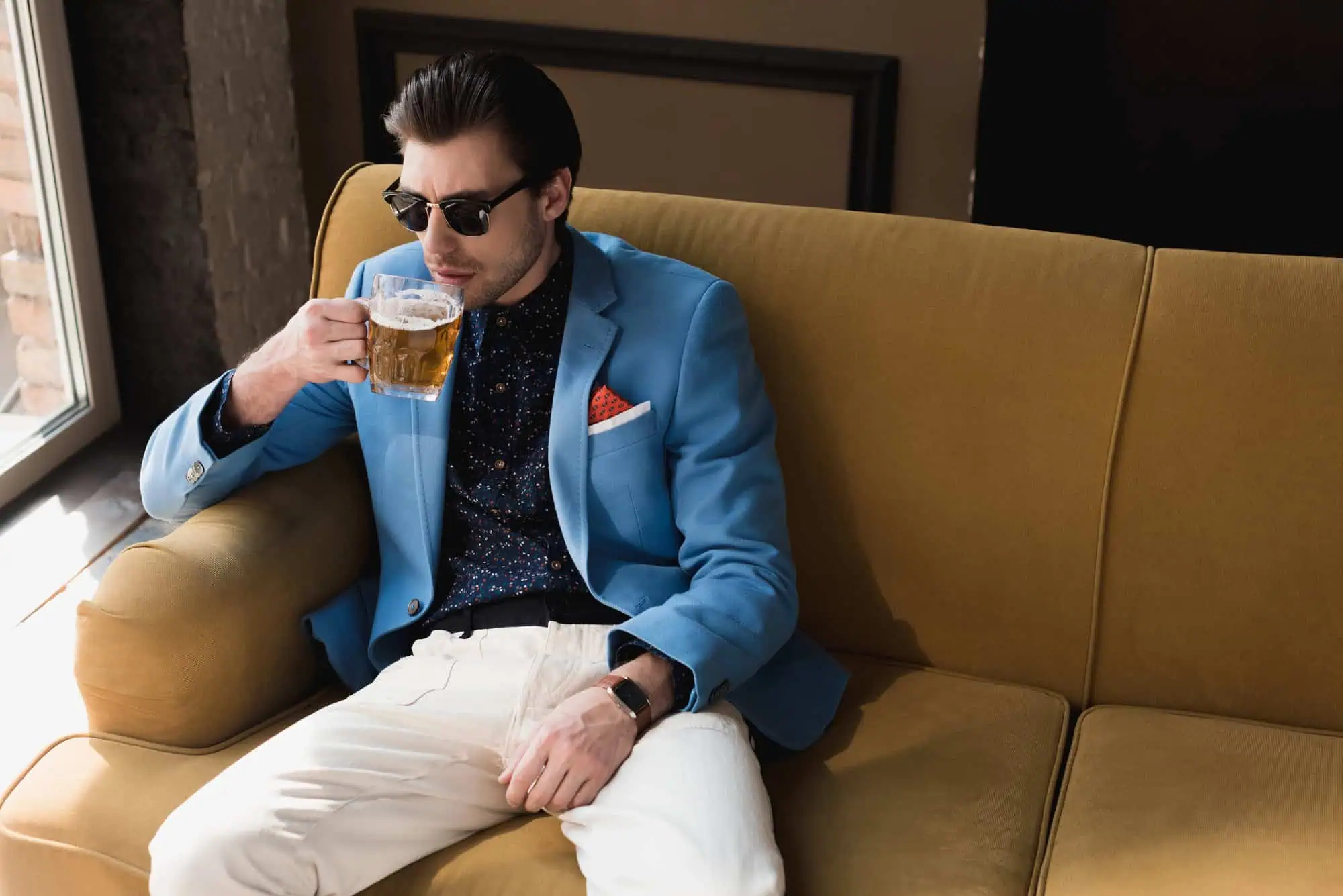 Attractive young man in stylish suit sitting on couch and drinking beer