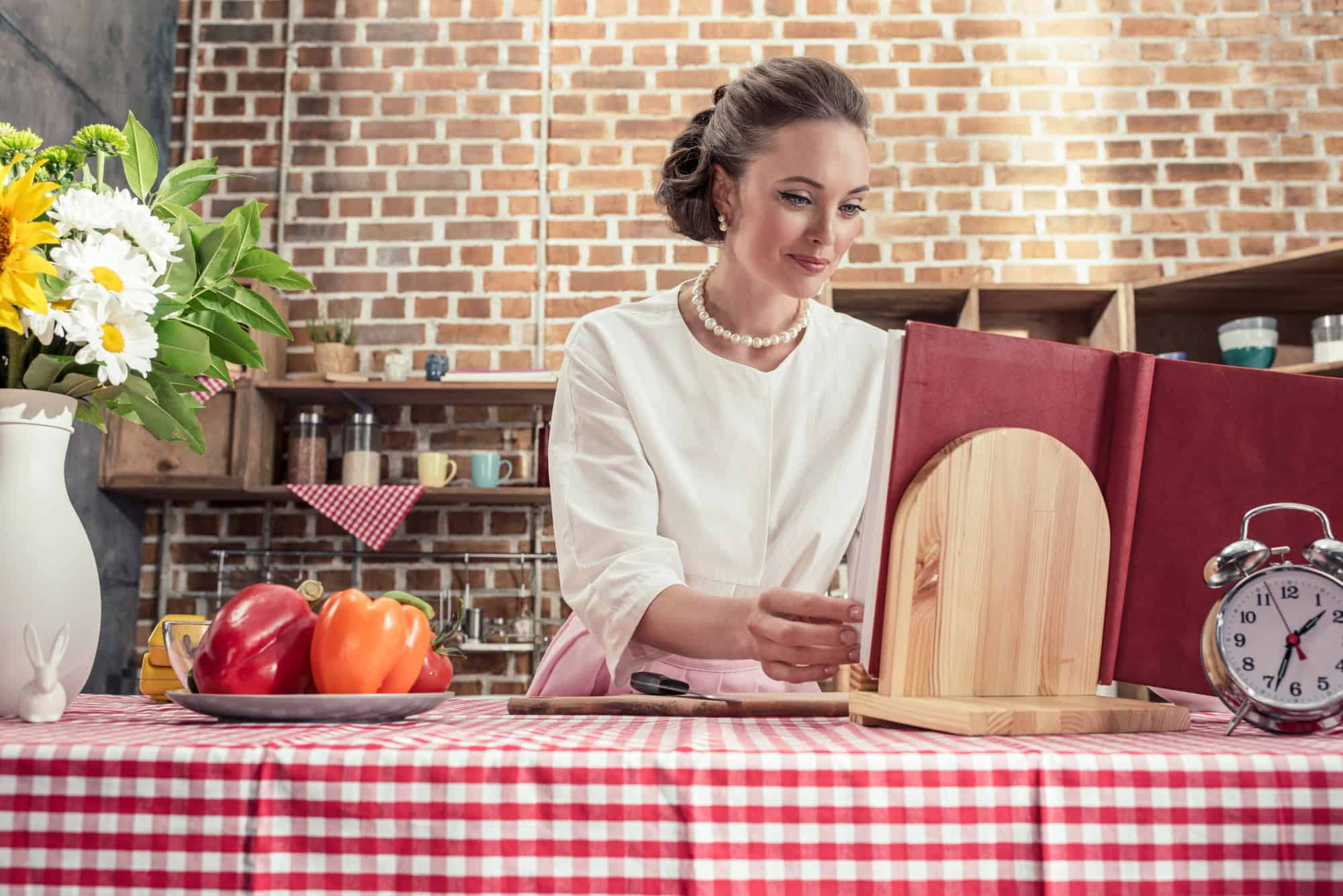Happy adult housewife in vintage clothes reading recipe book at kitchen
