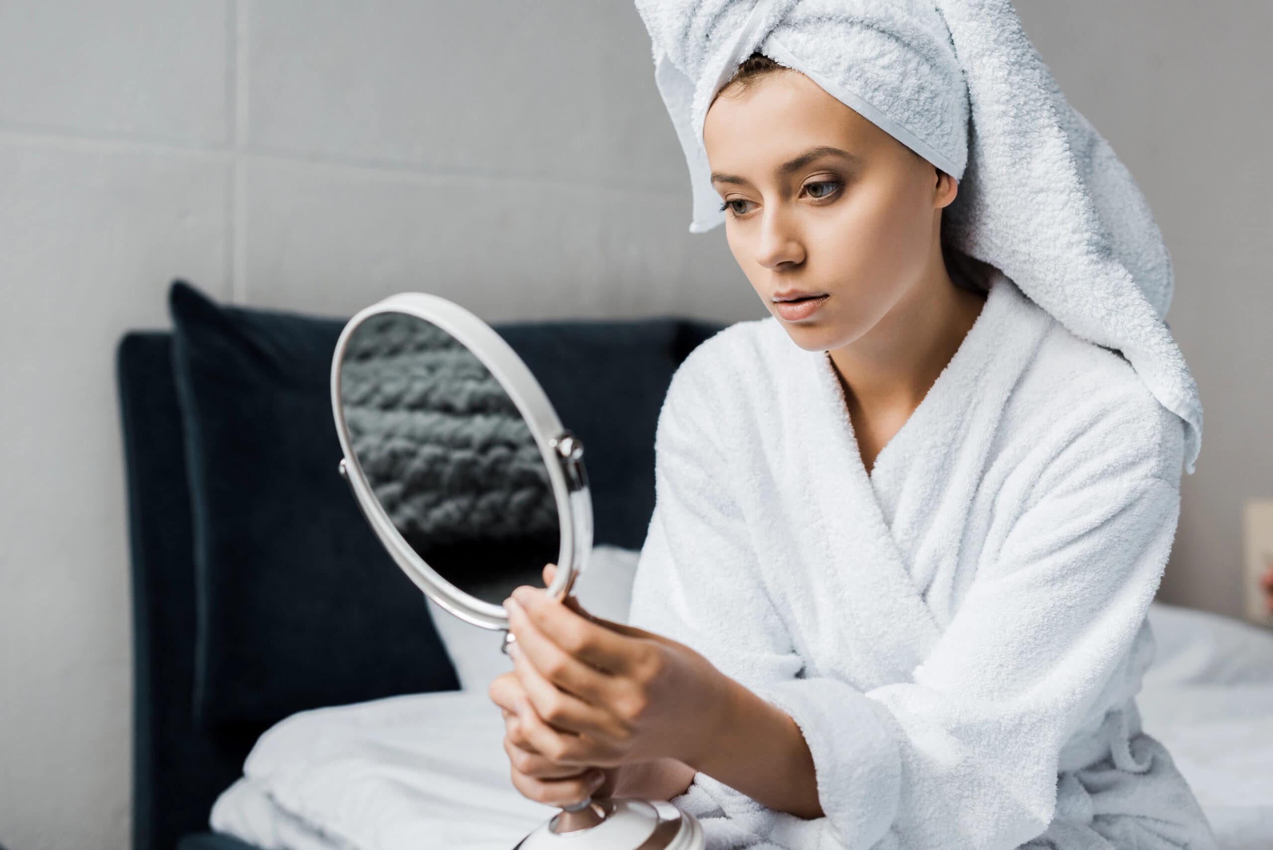 Attractive girl in white bathrobe and towel looking at her face in mirror