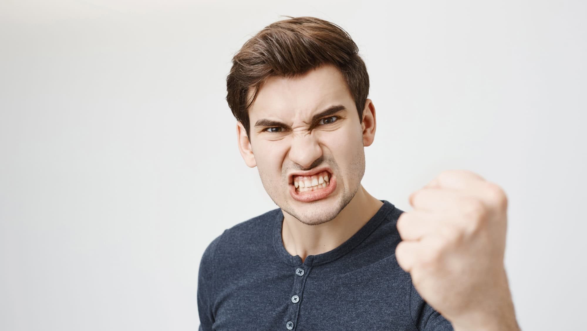 Fierce confident stylish european dark-haired male with trendy haircut holding fist in front of him as if ready for fight or challenge, clenching white teeth, having aggressive expression on face