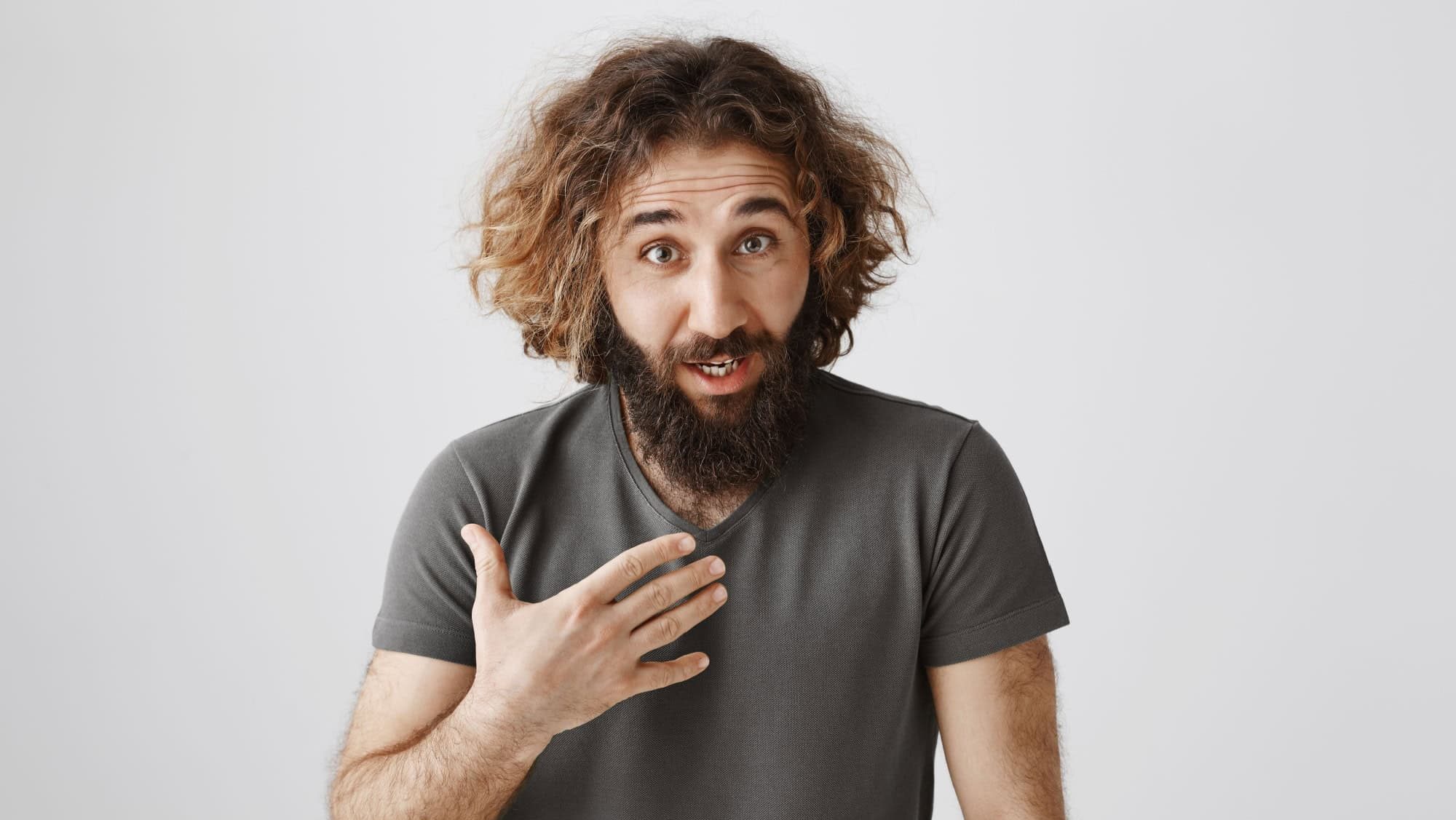 So it is my fault. Studio shot of displeased offended curly-haired male with beard pointing at chest and looking from under forehead with shocked and disbelieving expression, standing over gray wall
