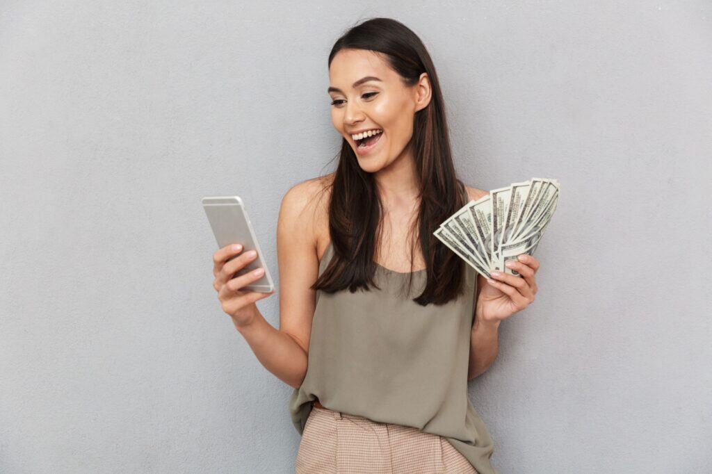 Portrait of a happy asian woman holding money banknotes and using mobile phone isolated over gray background
