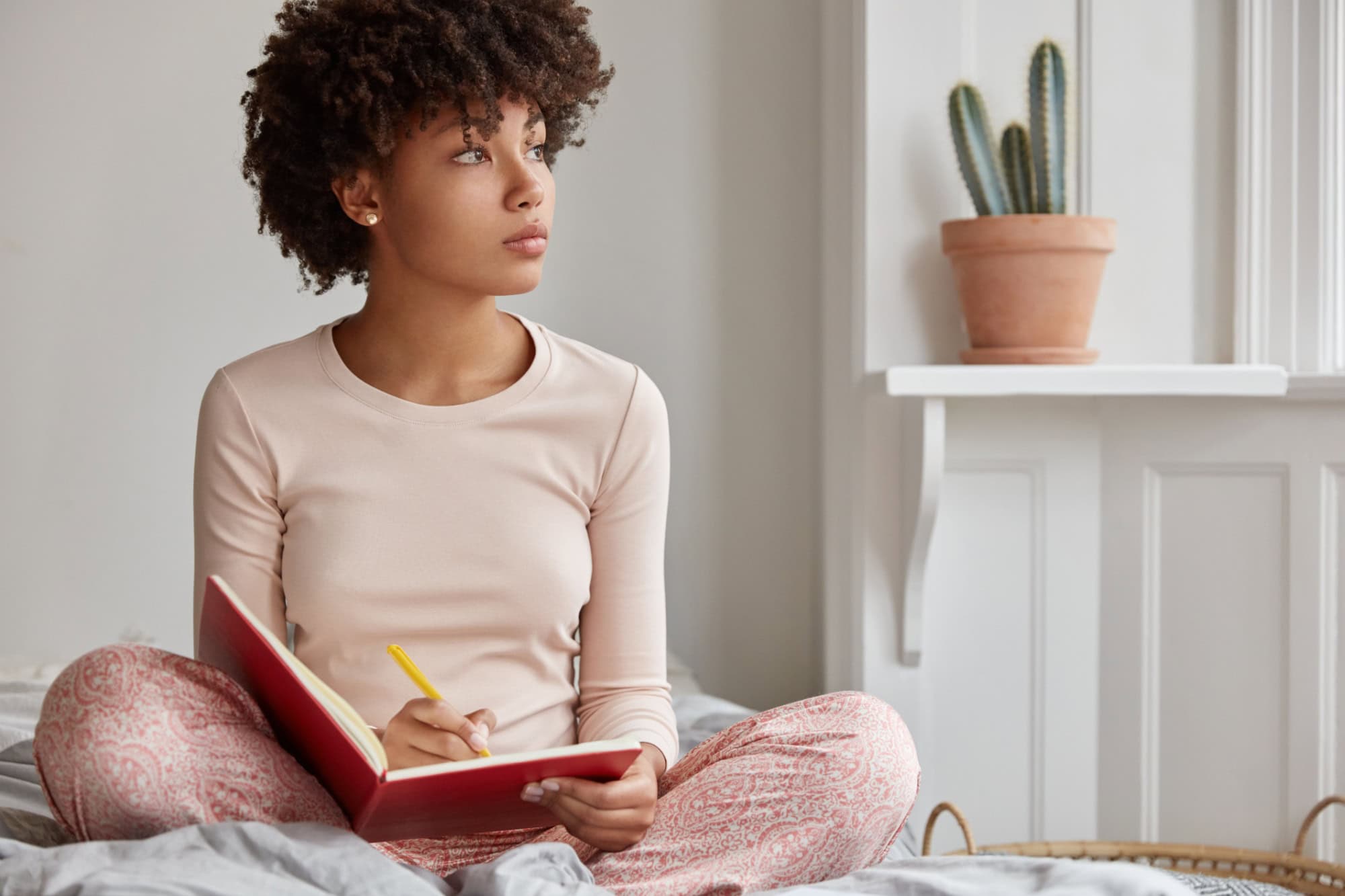 Cropped shot of dark skinned woman sits crossed legs, wears pyjamas, makes notes in diary, focused aside, blank copy space against domestic interior. Blogger creats publication for blog in notebook