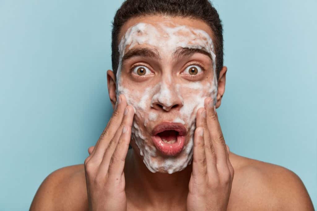 Portrait of surprised brown eyed man washes face with cleanser and soap, has foam on skin, being wet, stares in mirror, shocked to have acnes, isolated over blue background. Men hygiene