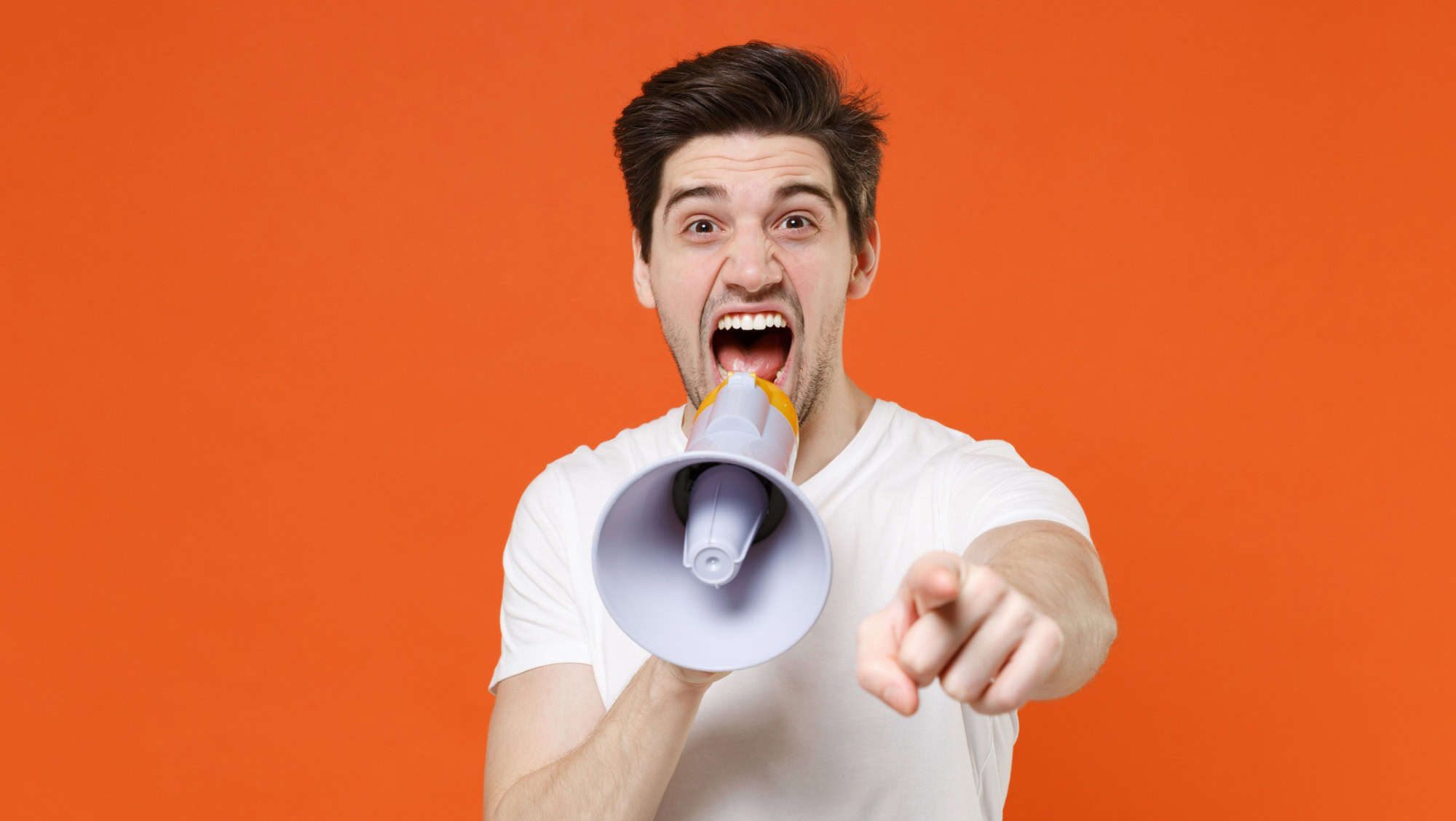 Crazy shocked young man 20s wearing basic casual empty blank white t-shirt standing screaming in megaphone pointing index finger on camera isolated on bright orange colour background studio portrait