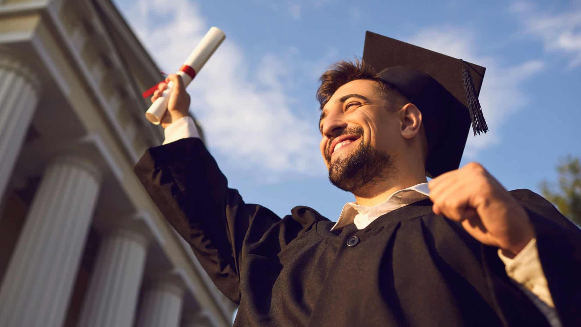 Low angle portrait of happy triumphant male graduate standing near university holding up diploma. From below of young handsome man proud of academic achievements celebrating college graduation