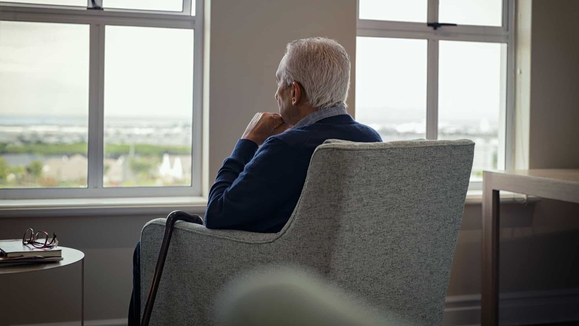Rear view of senior man sitting on armchair and looking through the window. Lonely old man sitting at home near window