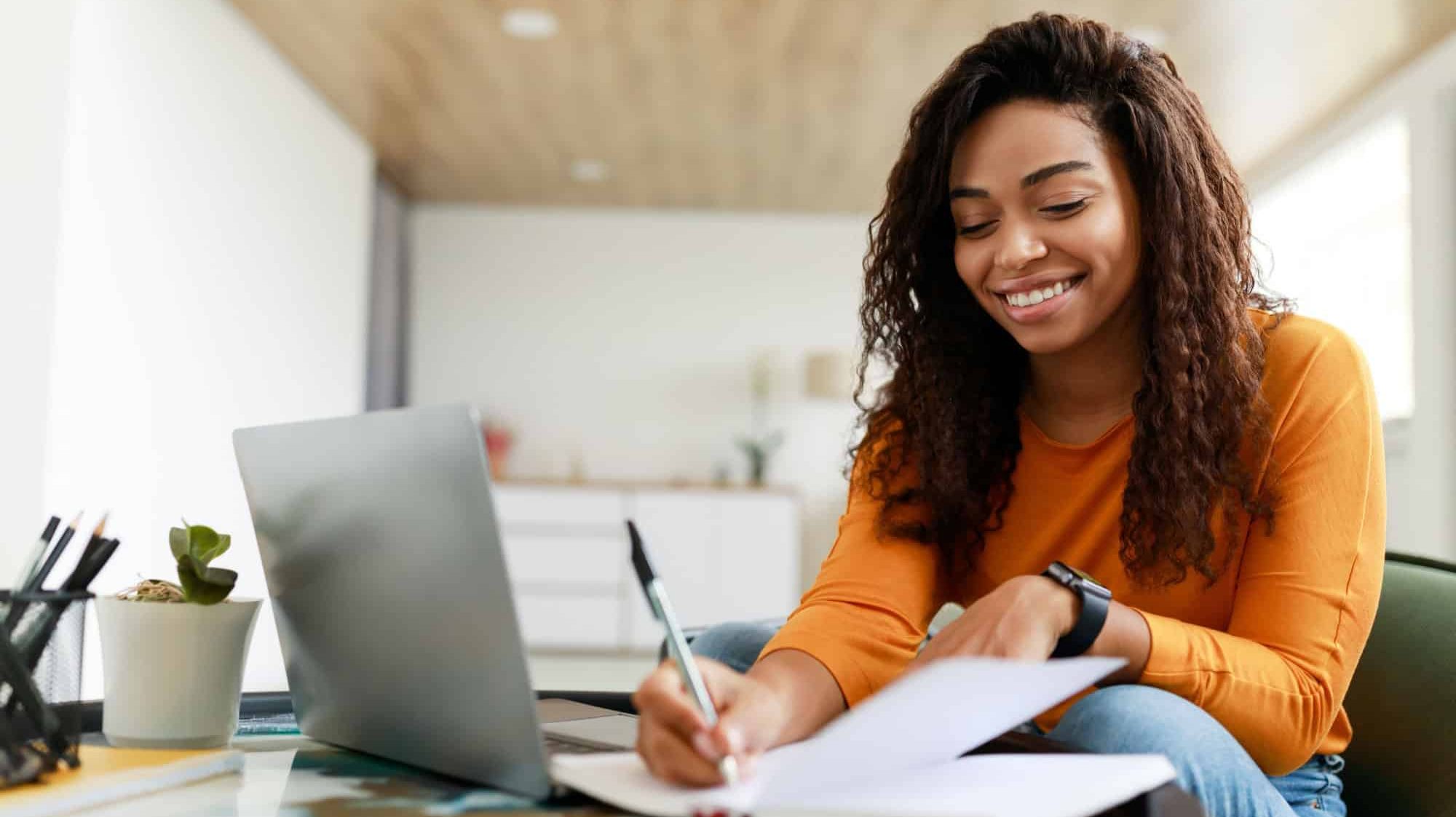 Business And Education Concept. Smiling young black woman sitting at desk working on laptop writing letter in paper notebook, free copy space. Happy millennial female studying using pc