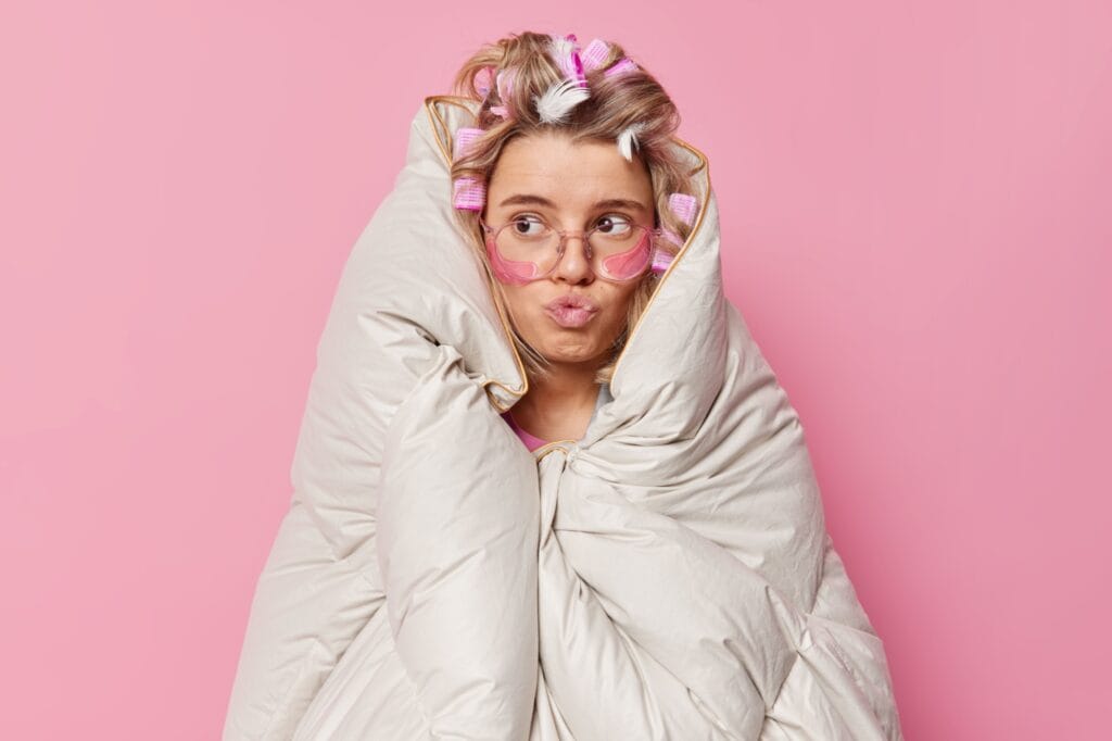 Surised funny woman pouts lips looks away wears big optical eyeglasses makes hairstyle applies rollers and hydrogel patches wrapped in blanket prepares for sleep isolated over pink background.