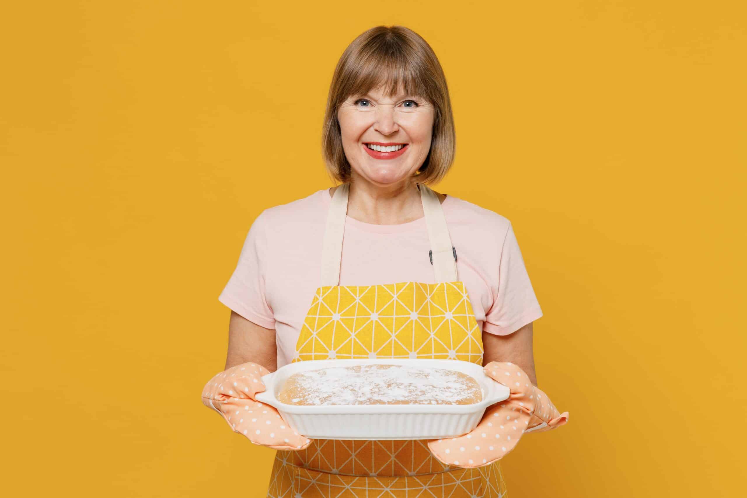 Elderly smiling happy fun cheerful housekeeper housewife woman 50s in orange apron hold casserole dinner meal isolated plain on yellow background studio portrait. People household lifestyle concept