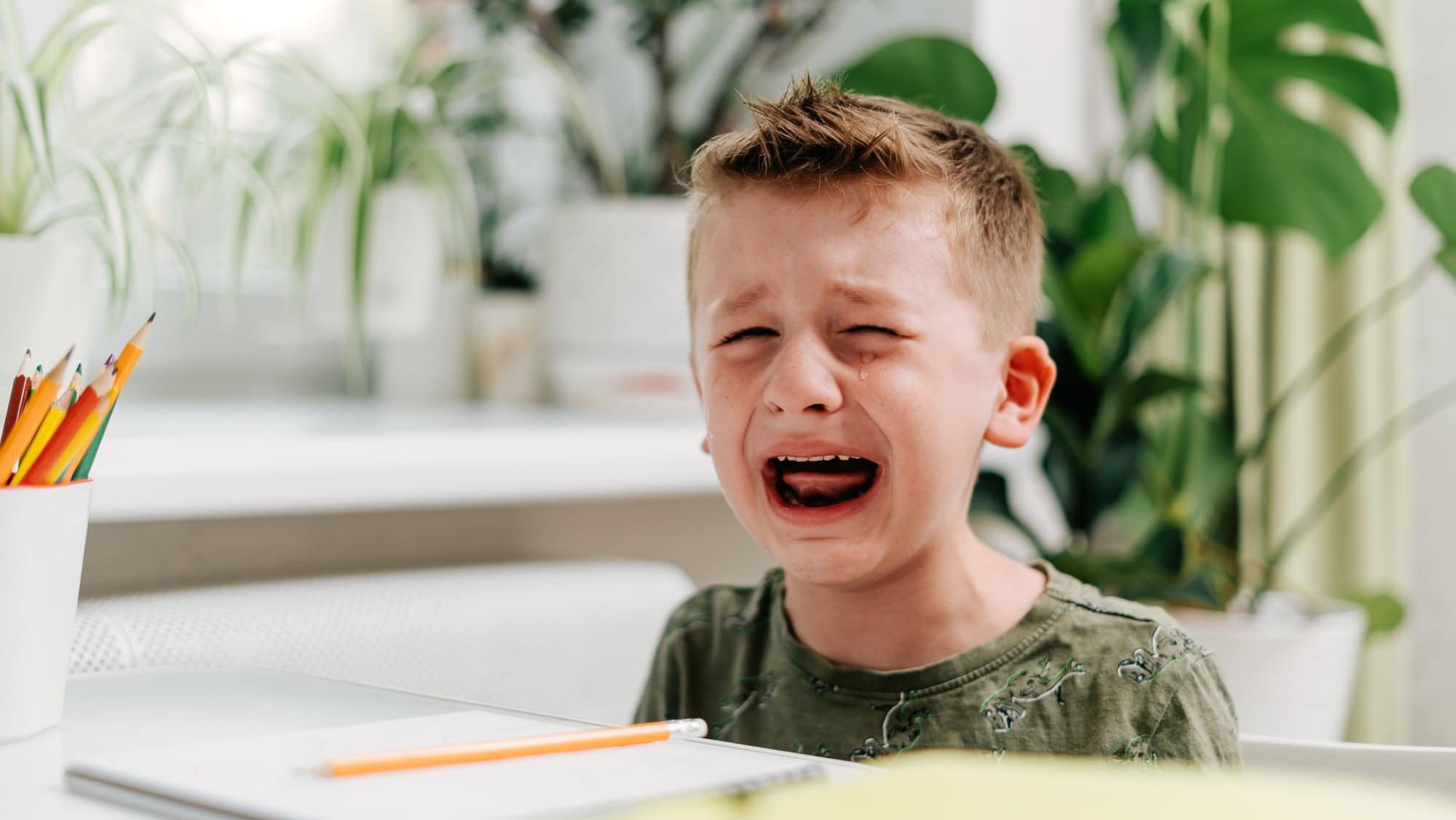 Back to school. Crying school kid boy studying at home and doing homework. Sad caucasian child siting at table with notepad, pencils and training books. Distance learning online education.