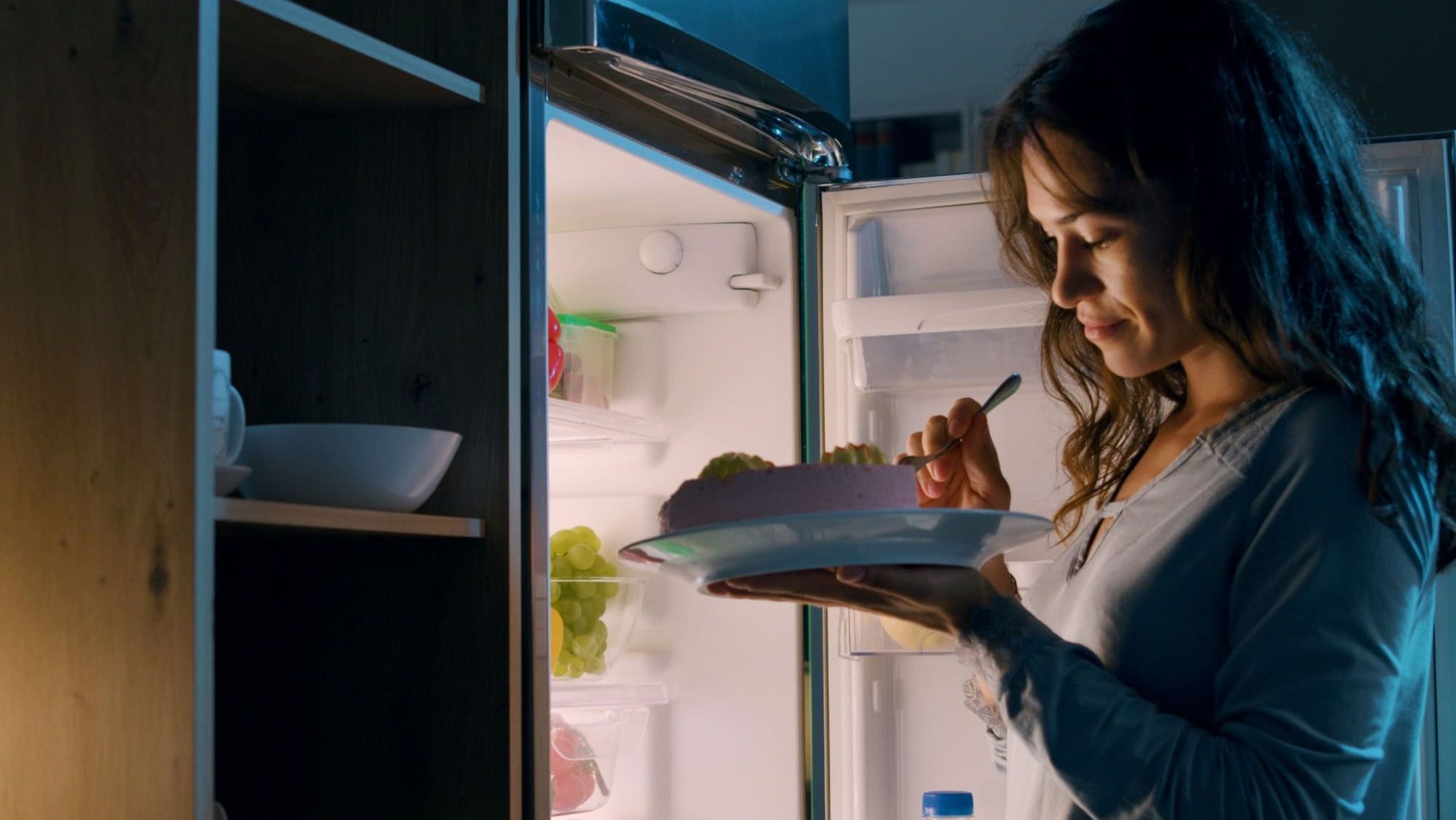 Happy hungry woman having a midnight snack, she is standing in front of the fridge and eating a delicious cake