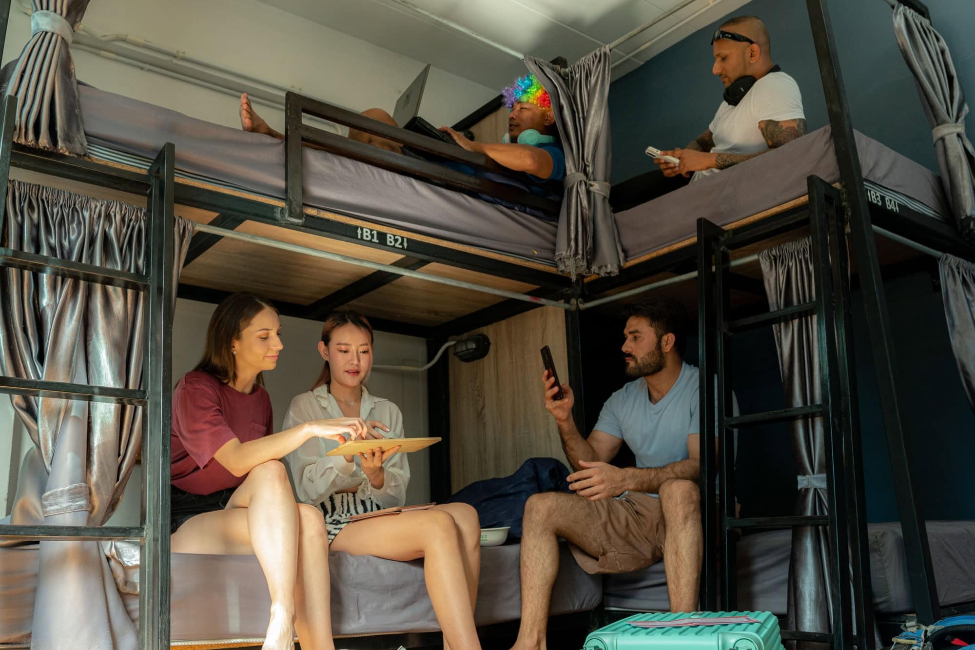 Groups of travelers from various nationalities gather in a hostel, selfies to keep memories, funny man using smartphone in upper bed, backpackers.