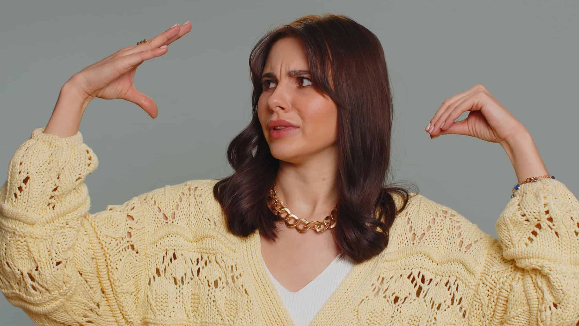 Bored pretty young woman in cardigan showing blah blah gesture with hands, has not interested in the conversation, gossips, empty promises, lier. Millennial girl isolated on gray studio background