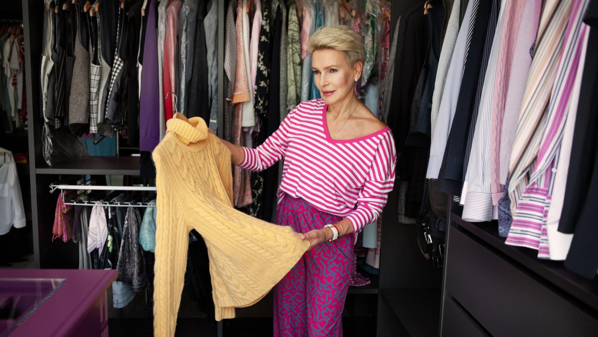 Mature Woman organizes or chooses clothes outfit and tries on different color of sweater in large wardrobe closet at home.