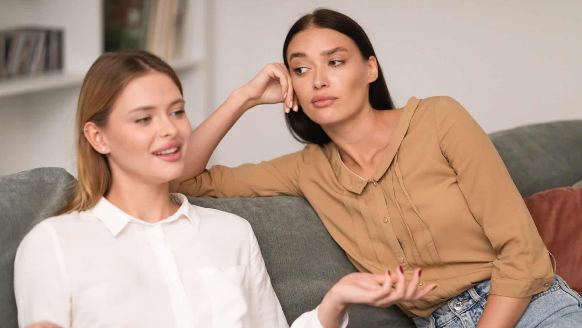 Jealous Woman Listening With Envy To Her Friend Bragging About Great Life Sitting On Sofa In Modern Living Room At Home. Envious Lousy Friends, Fake Friendship Concept. Selective Focus