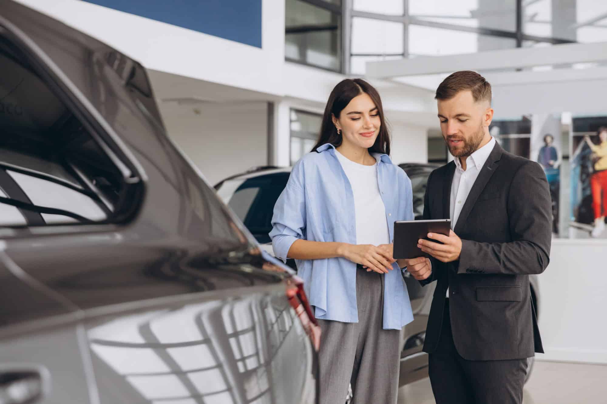 Young woman customer female buyer client wears shirt consult with salesman hold tablet choosing auto want to buy new automobile in car showroom vehicle salon dealership store motor show.
