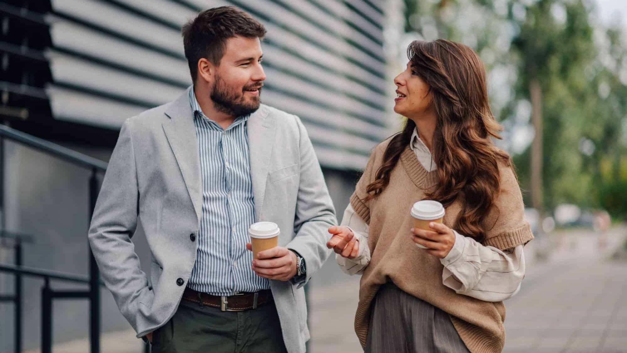 Two business people walking and talking outdoors, holding coffee cups, dressed in modern attire, happy and confident during a break from the office. Enjoying beverages on a city street in autumn