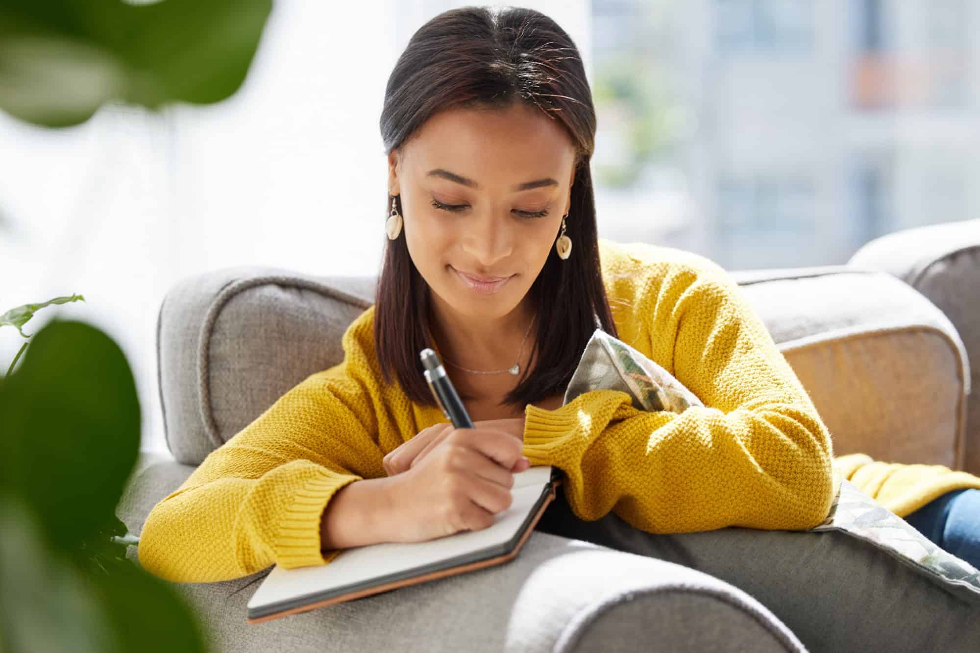 Woman, relax and writing in journal at house for diary entry, personal growth and wellness affirmations. Female person, book and creativity for goals reminder, inspiration quotes and gratitude letter