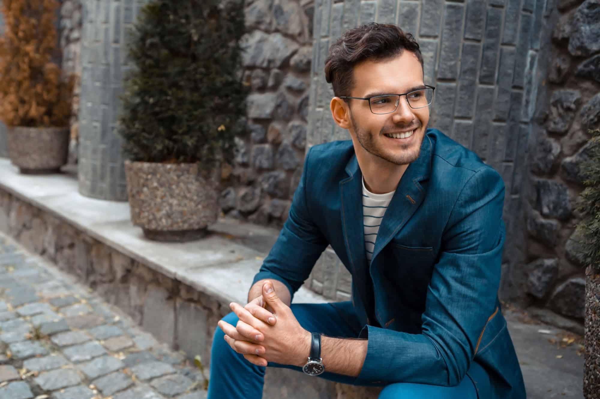 Portrait of stylish handsome young man with bristle sitting on parapet outdoors. Man wearing jacket and shirt
