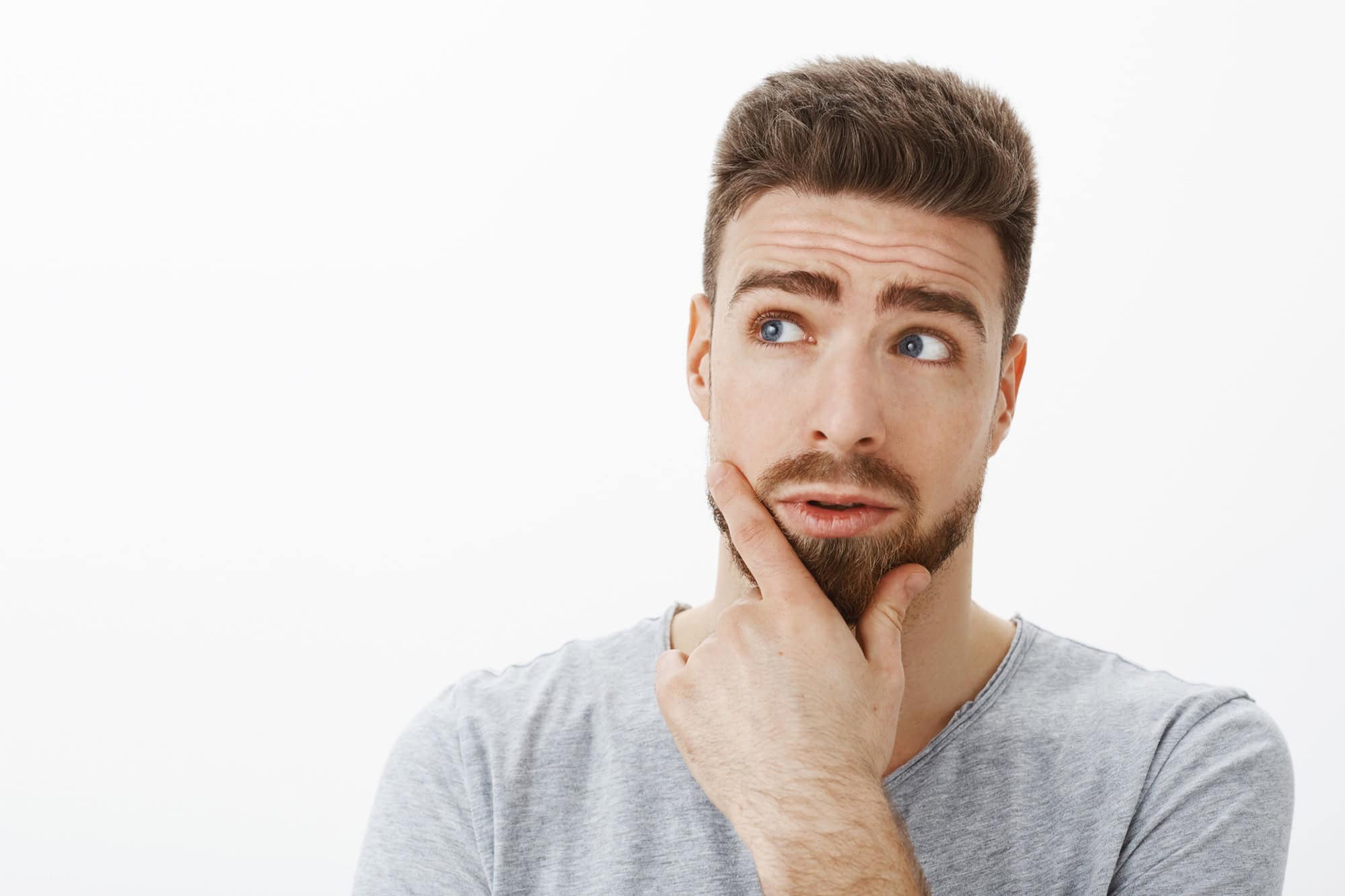 Curious and questioned handsome businessman trying solve problem thinking standing in thoughtful pose rubbing beard looking at upper left corner making decision against gray background
