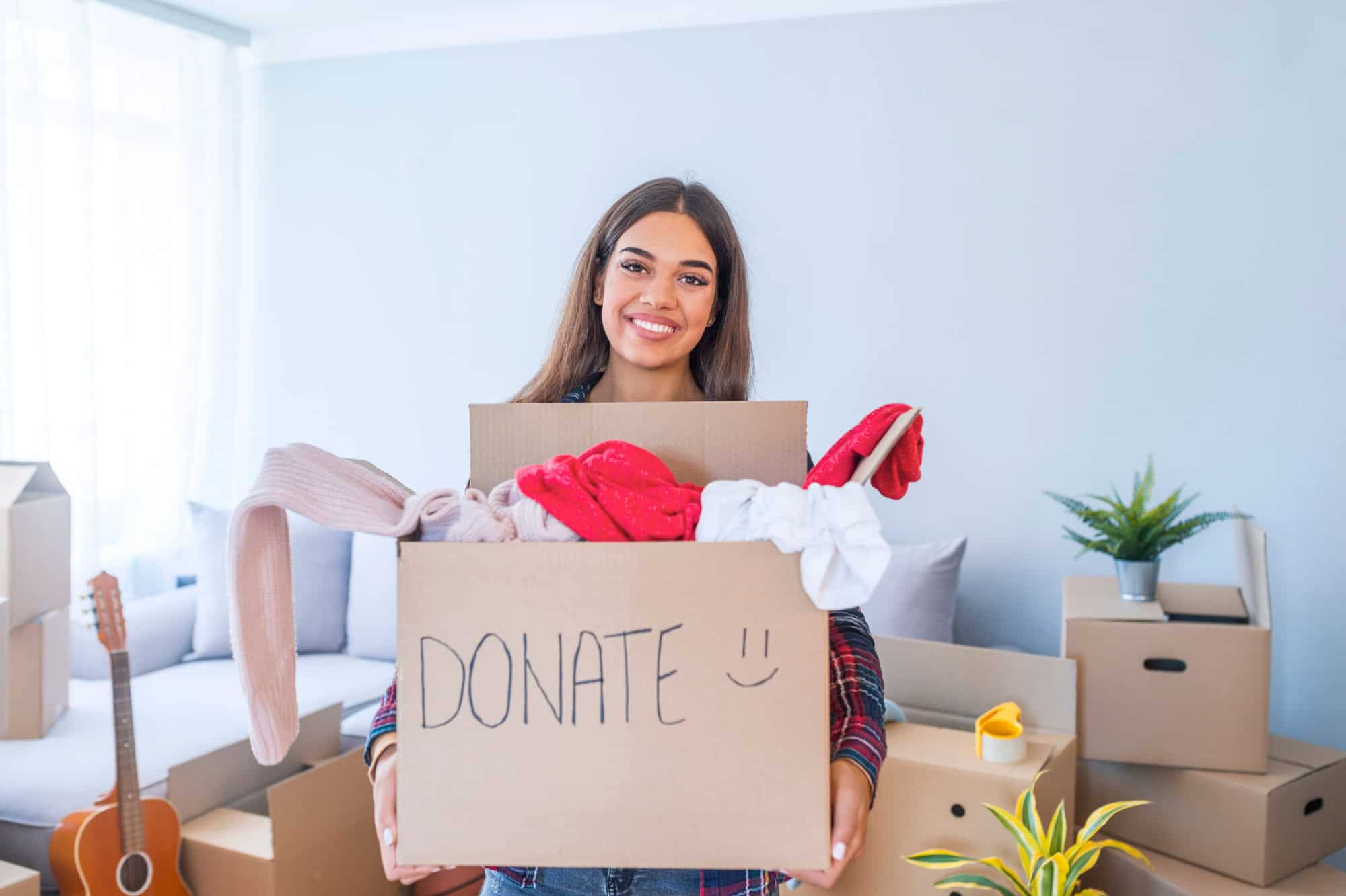 Young woman with donation box at home. Donation box for poor with clothing in female hands. Woman donates clothing to shelter. Cheerful woman holds box of donated clothing