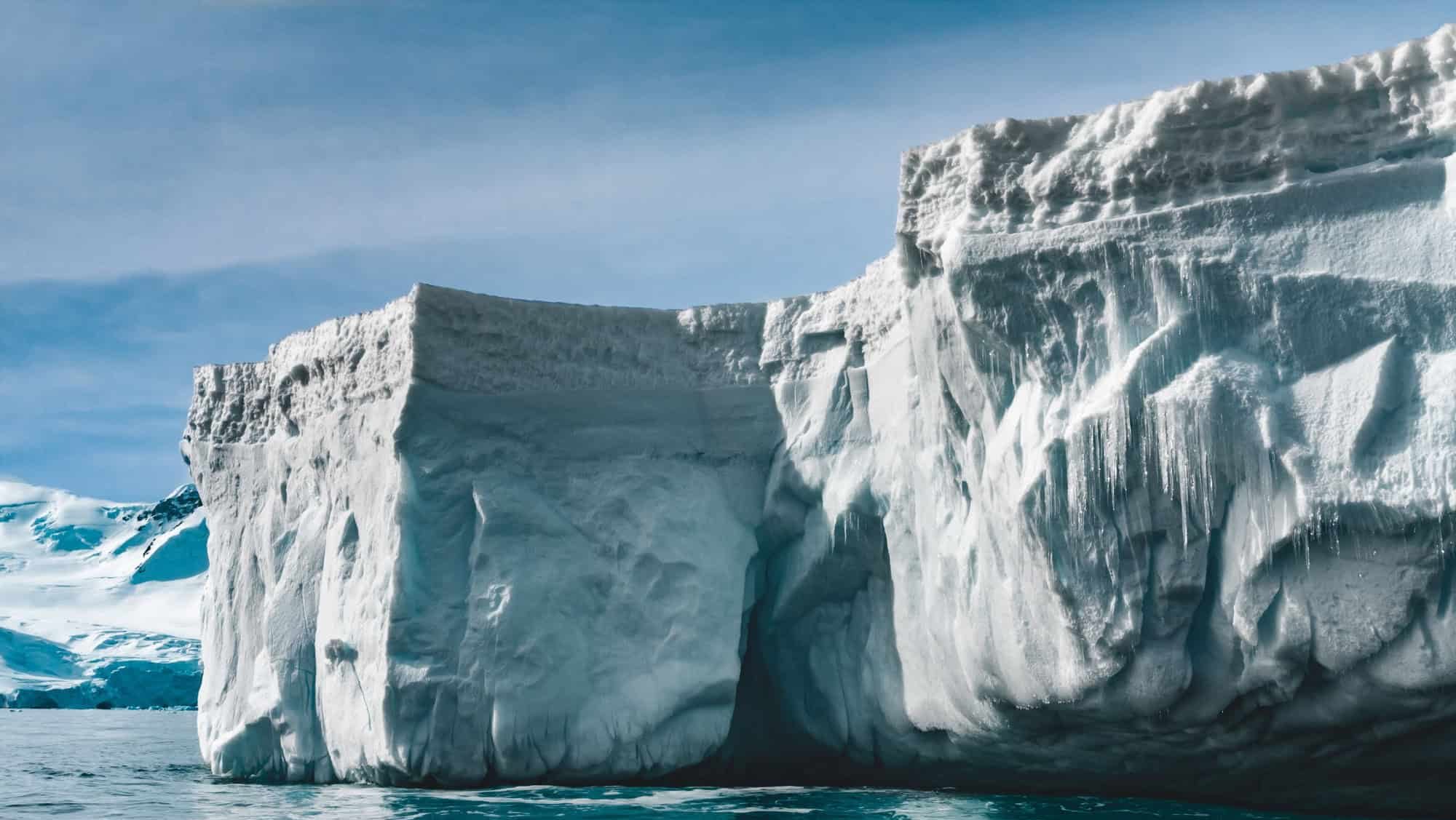 Close-up sunlit iceberg. Antarctica scene in blue and white tints. Amazing snow covered block of ice with icicles floating among the polar ocean. The cloudy sky background. Picturesque winter scenery.