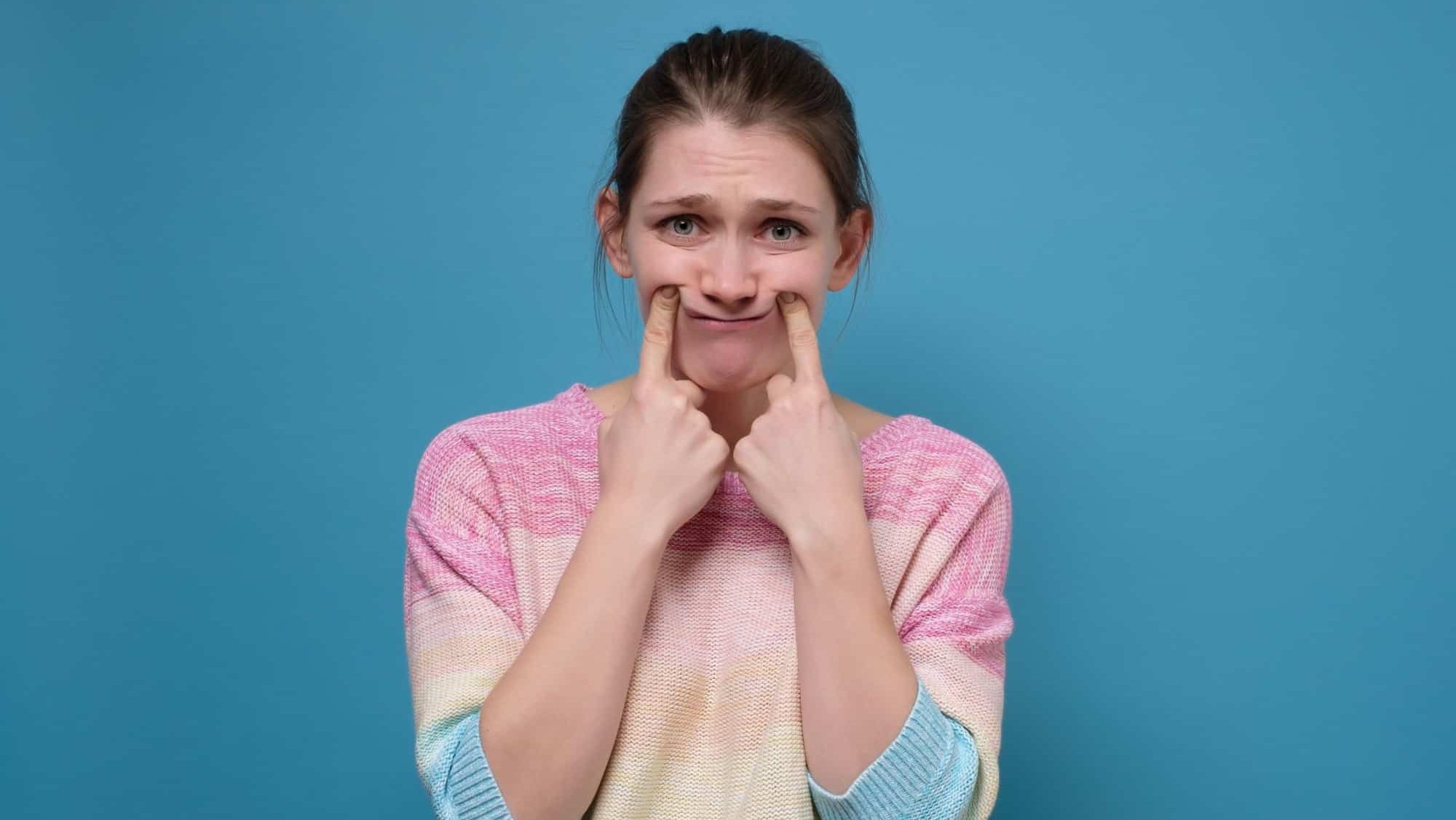 Young upset woman making fake smile with her fingers stretching the corners of her mouth. Try to stay positive after failing at something.