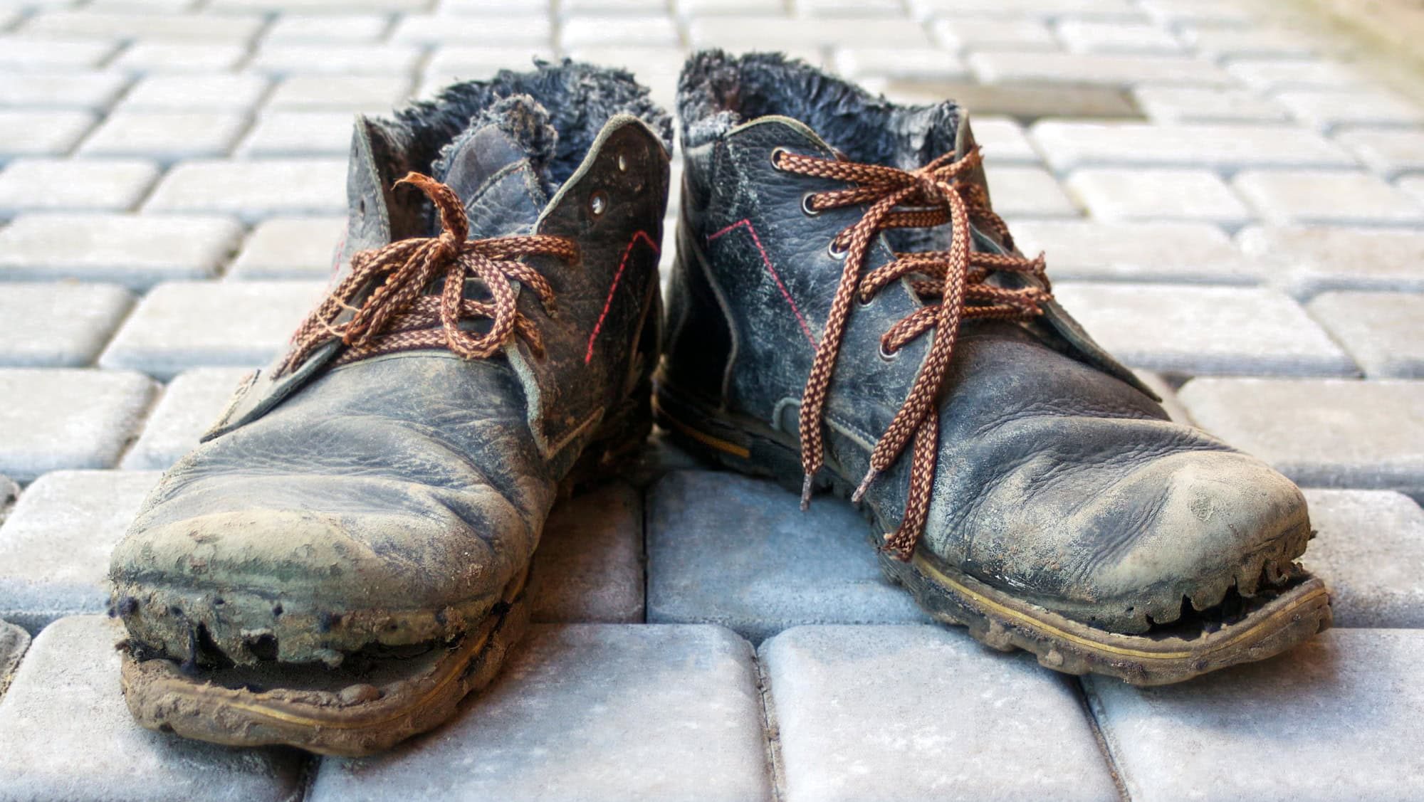 A pair of dirty boots. Old worn dark blue leather shoes with variegated brown laces. Background from gray pavers. The concept of poverty, homelessness, lack of money. Selective focus