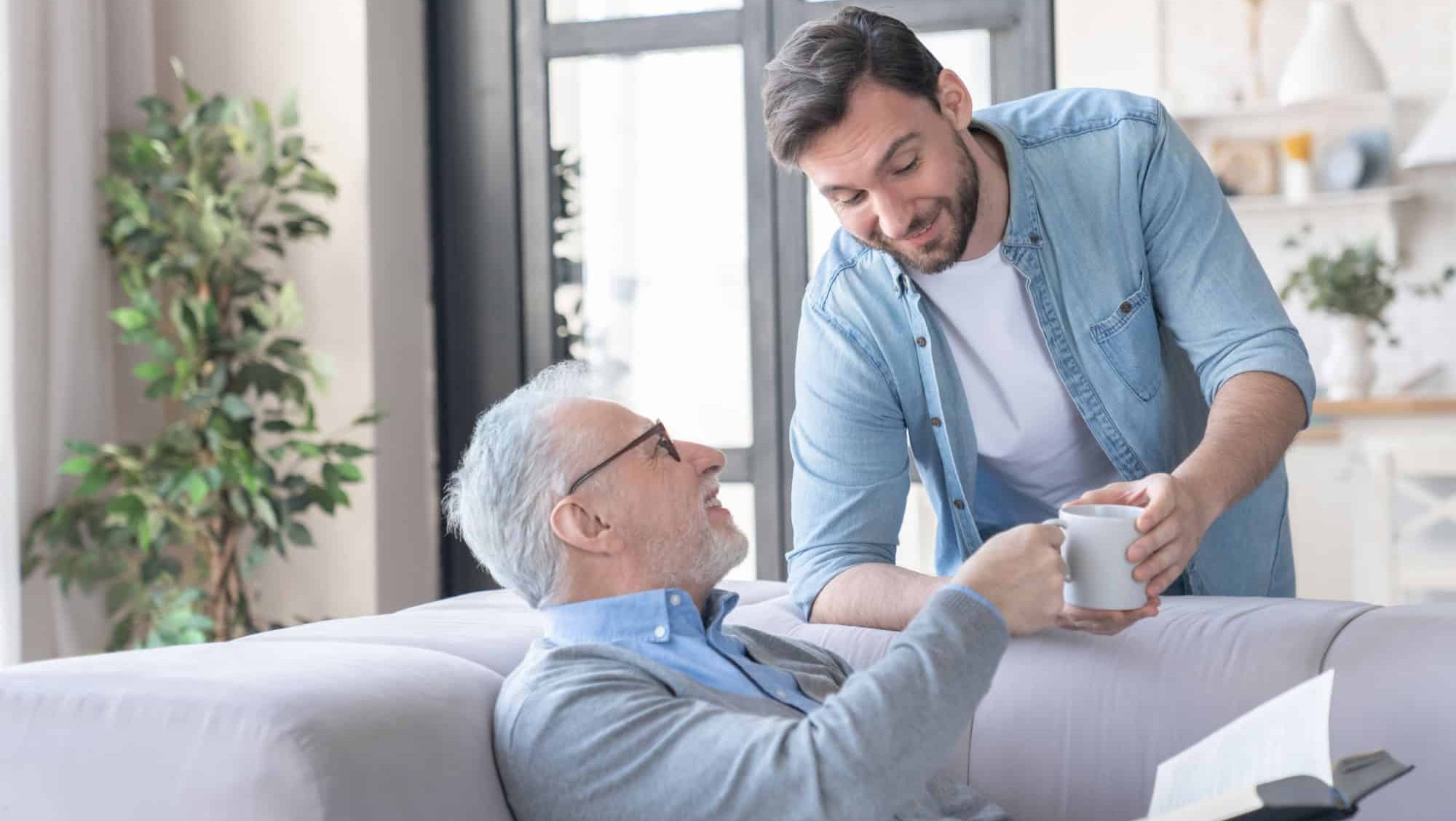 Loving caring young adult son bringing his old elderly senior father a cup of water hot drink at home indoors. Elderly generation support and help aid. Happy father`s day! I love you, dad!