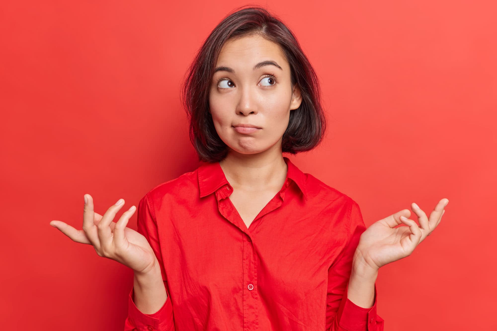 Clueless hesitant beautiful Asian woman with short dark hair spreads palms sideways shrugs shoulders has indecisive expression dressed in shirt stands against vivid red background. Who knows.