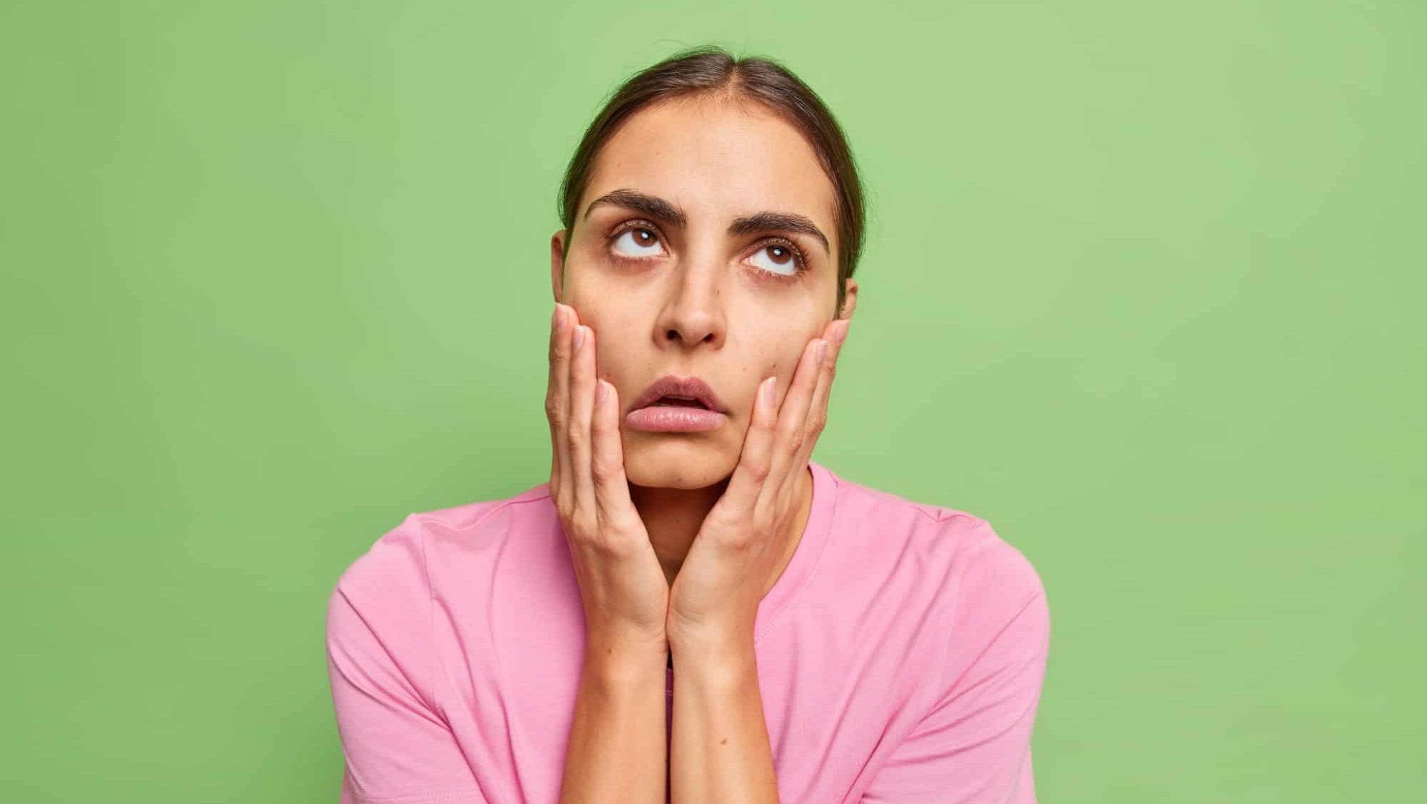 Uninterested bored young woman keeps hands on cheeks fed up of dull conversation looks above makes facepalm and rolls eyes hears nonsense wears casual t shirt isolated over green background.