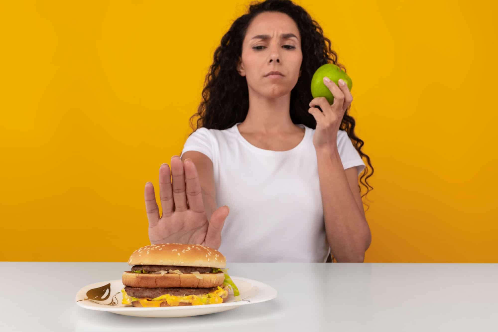 No Junk Food. Serious Lady Refusing To Eat Unhealthy Burger Choosing Apple, Showing Stop Sign Gesture With Palm Sitting At Table, Selective Focus On Cheeseburger And Hand. Healthy Eating, Diet Concept